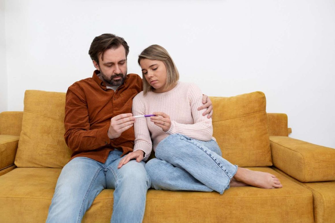 medium-shot-couple-sitting-couch (1) A man and woman sit on a couch, both looking at a thermometer with concerned expressions.
