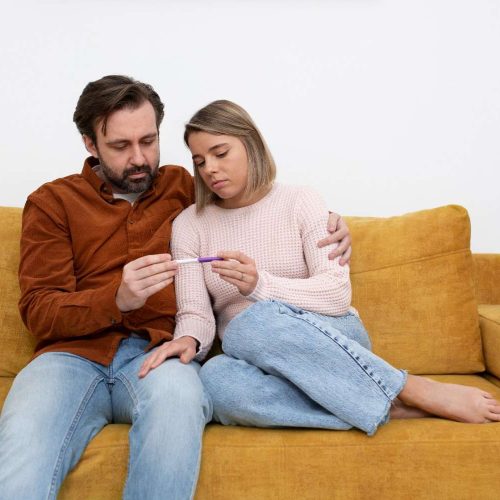 A man and woman sit on a couch, both looking at a thermometer with concerned expressions.
