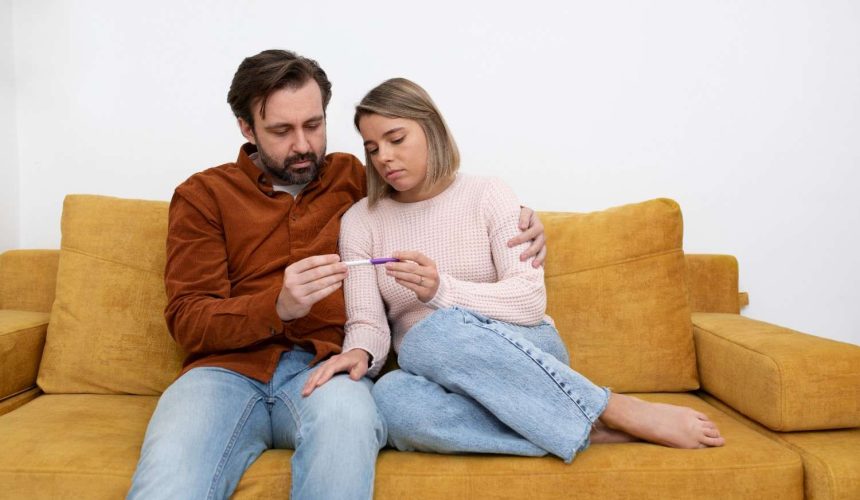 medium-shot-couple-sitting-couch (1) A man and woman sit on a couch, both looking at a thermometer with concerned expressions.