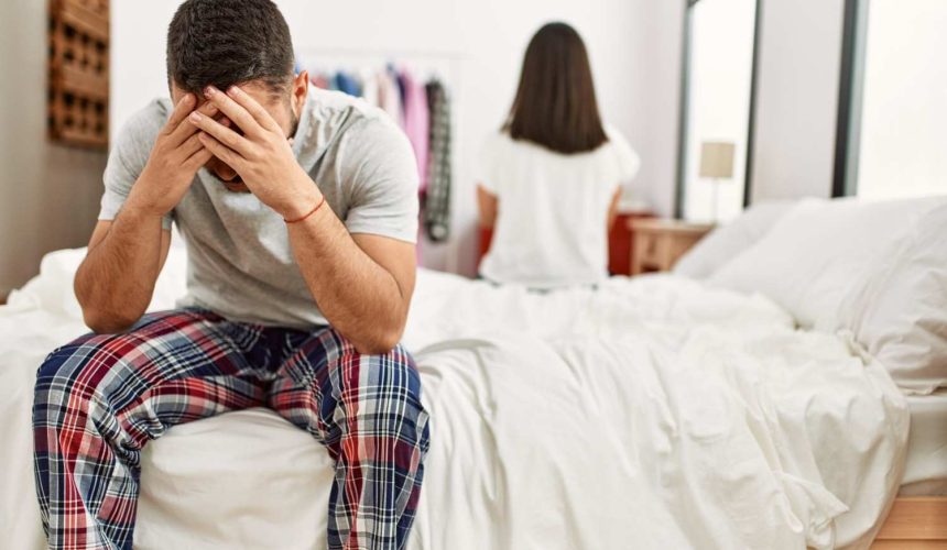 young-latin-couple-trouble-sitting-bed-with-hands-head (1) A man is seated on a bed, with his head in his hands, expressing feelings of sadness or frustration.